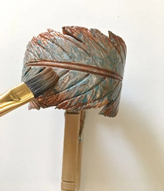 step 25 - When the bracelet has cooled, brush it with the floor polish to give it a sheen and protect the baked on mica powder. Use gentle, quick strokes working from the center of the feather out to the edges. Clean the brush on a paper towel between strokes to avoid smearing the colorful clay with excess loose powder that might remain on the surface. Attach a clothespin to keep the bracelet from rolling around as you work.