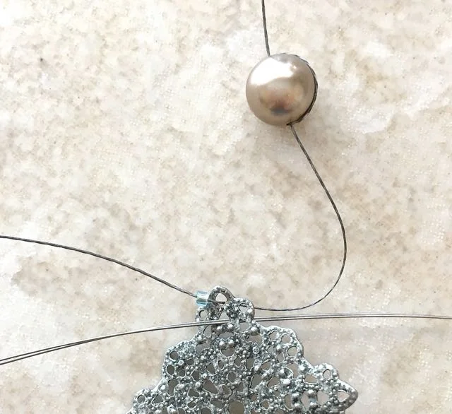 step 3 - Pass the needle through the top hole of the component. Add a seed bead to the thread. Bring the needle through a hole below the first insertion point from front to back.