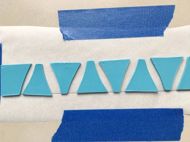step 9 - Attach an 8” x 2” piece of parchment paper to a ceramic tile with painters tape. Draw a straight line horizontally across the paper. Place the turquoise clay pieces on the line with a little spacing between them. This doesn’t need to be precise, as you will be adjusting them later.