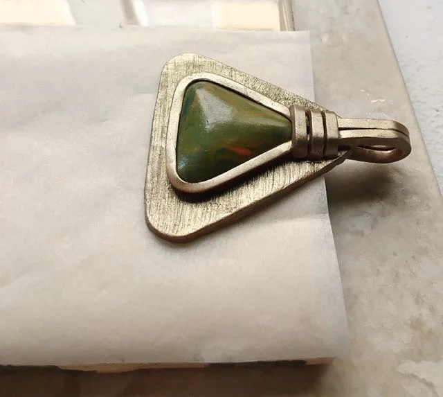 step 21 - Lay the pendant on parchment supported by a ceramic tile. Allow the upper end of the project to extend off the tile as shown to create a flat pendant. If the pendant is placed in the center of the tile, the thickness on the back of the bail will prevent the pendant from remaining completely flat.