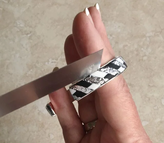 step 13 - Carefully peel off the film. Avoid handling the clay too much by slipping your fingers and spreading them inside the cuff to hold it. Press a blade against the inside edge of the bracelet channel on each side to remove excess clay and create even edges. Pressing the blade down is safer than dragging, which may tear the silver leaf. A crackled effect is nice but you don’t want to rip off large chunks of the silver.