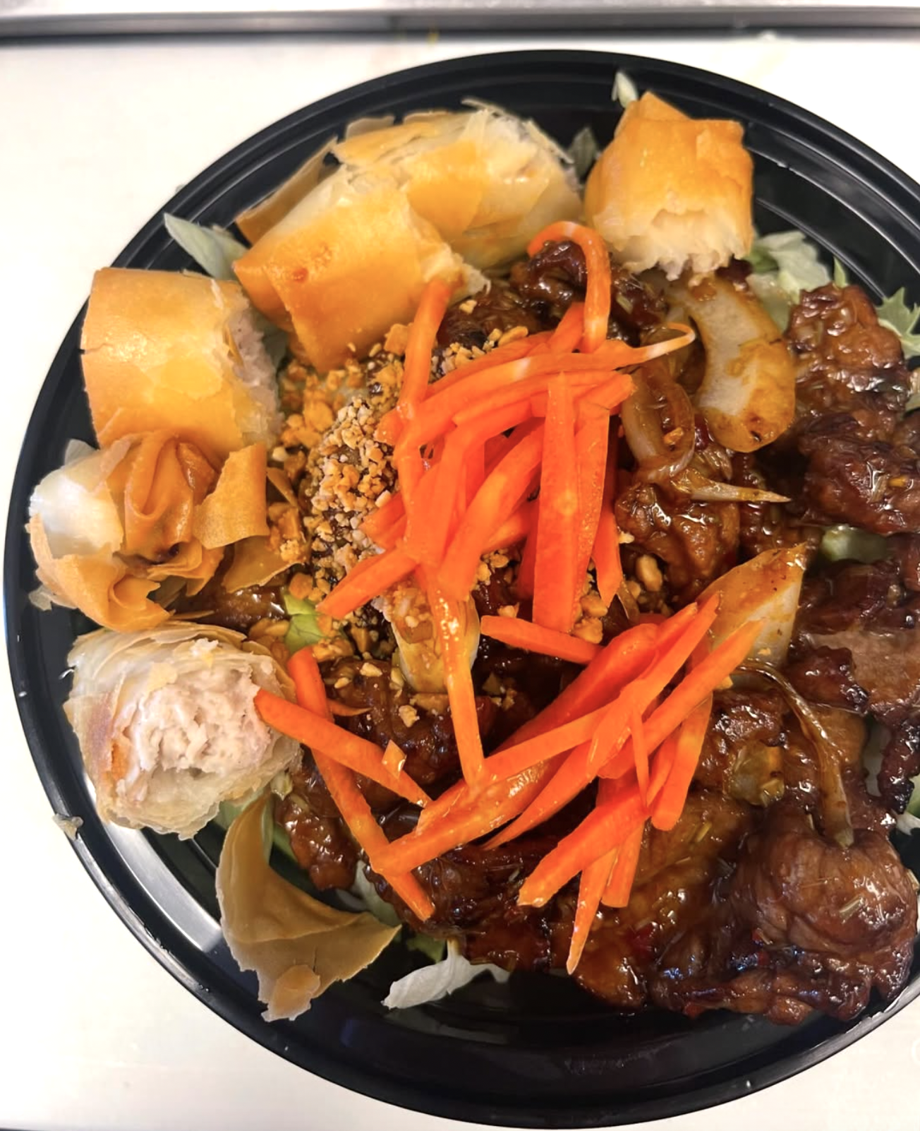 A bowl of Asian-style food with dumplings, shredded carrots, chopped peanuts, beef with sauce, and spring rolls.