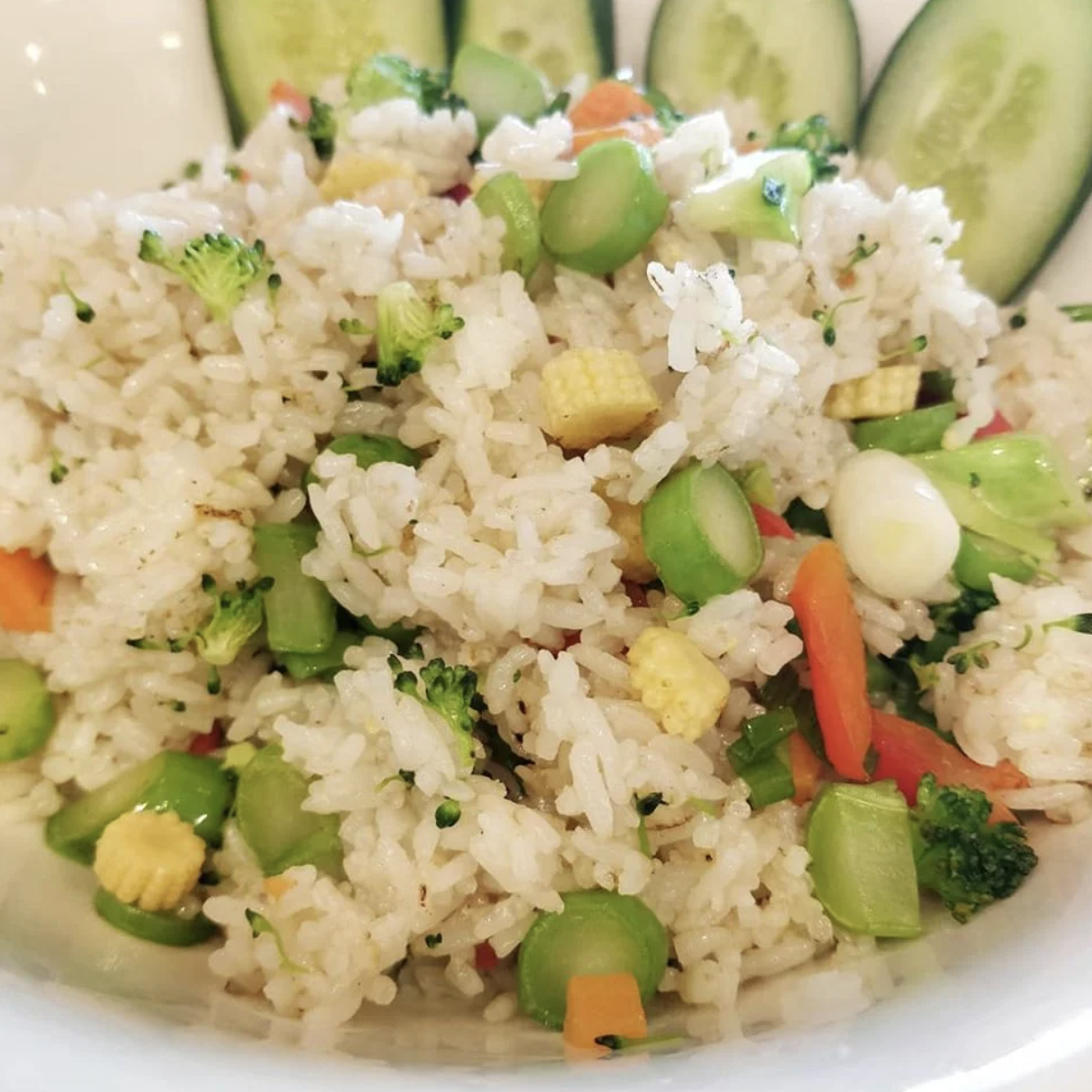 Close-up of a bowl of vegetable fried rice with green peas, broccoli, carrots, baby corn, and sliced cucumber garnished with cucumber slices in the background.