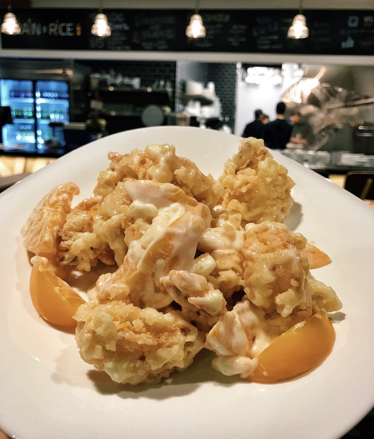 Plate of fried chicken pieces with creamy sauce and orange wedges in a restaurant with people working in the kitchen in the background.