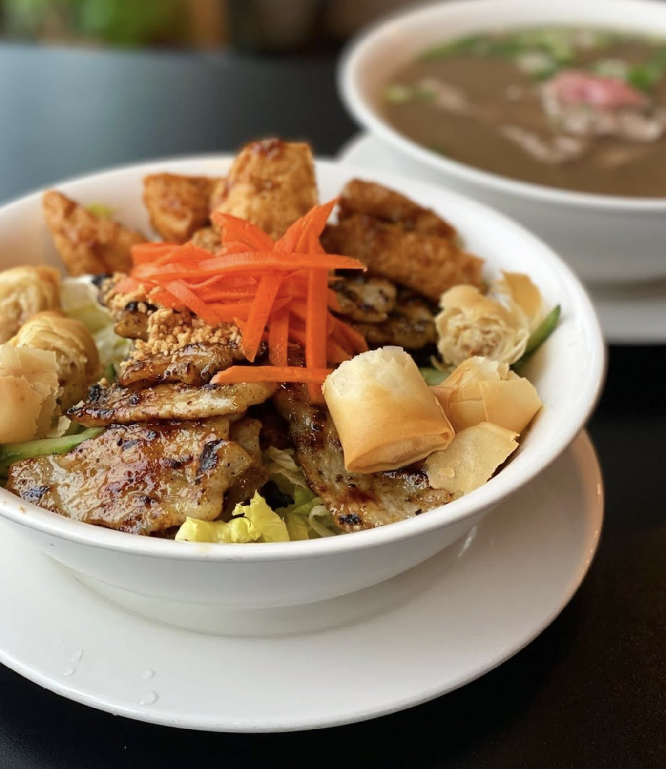 A white bowl filled with Asian-inspired grilled chicken, shredded carrots, fried crispy rolls, and spring rolls, with a bowl of hot soup in the background.