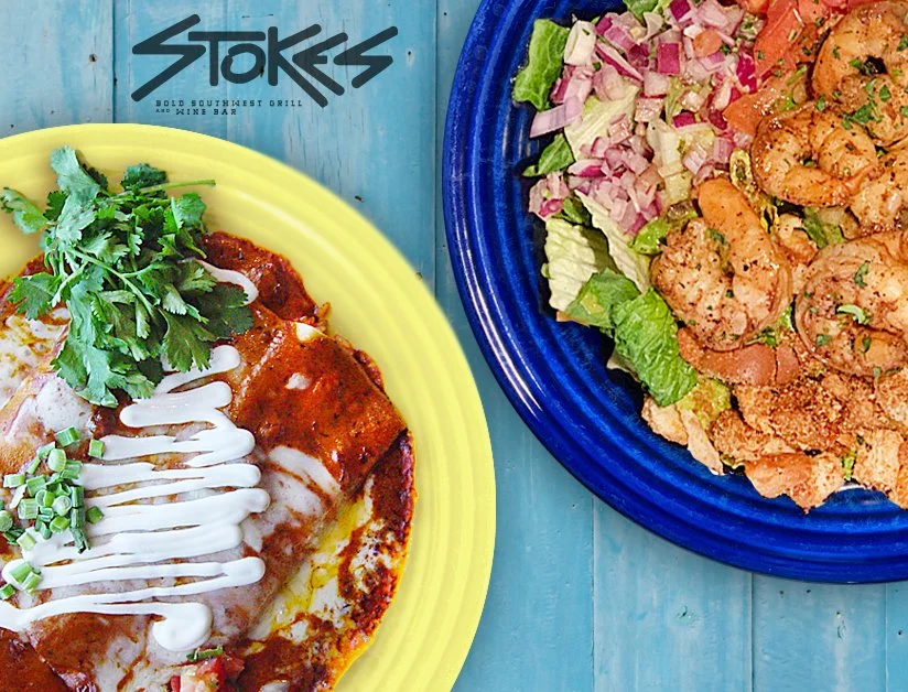 Blue wooden table with a yellow plate featuring enchiladas covered in red sauce and drizzled with sour cream. There is also a blue plate featuring a grilled shrimp salad with chopped tomatoes and red onion.