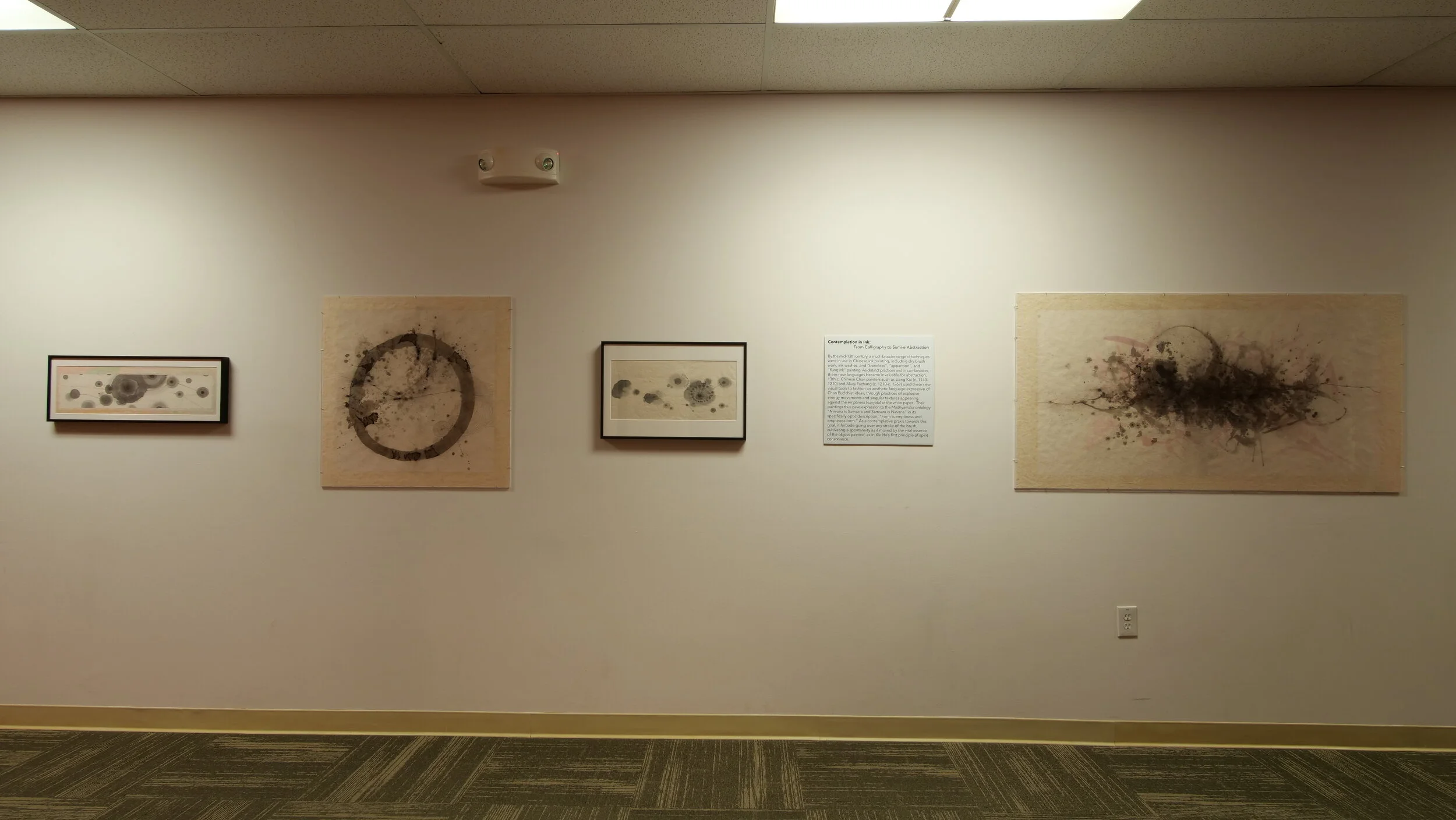 View of a long wall featuring abstract ink work in black and soft colors on canvas and other media of varied shapes and sizes.