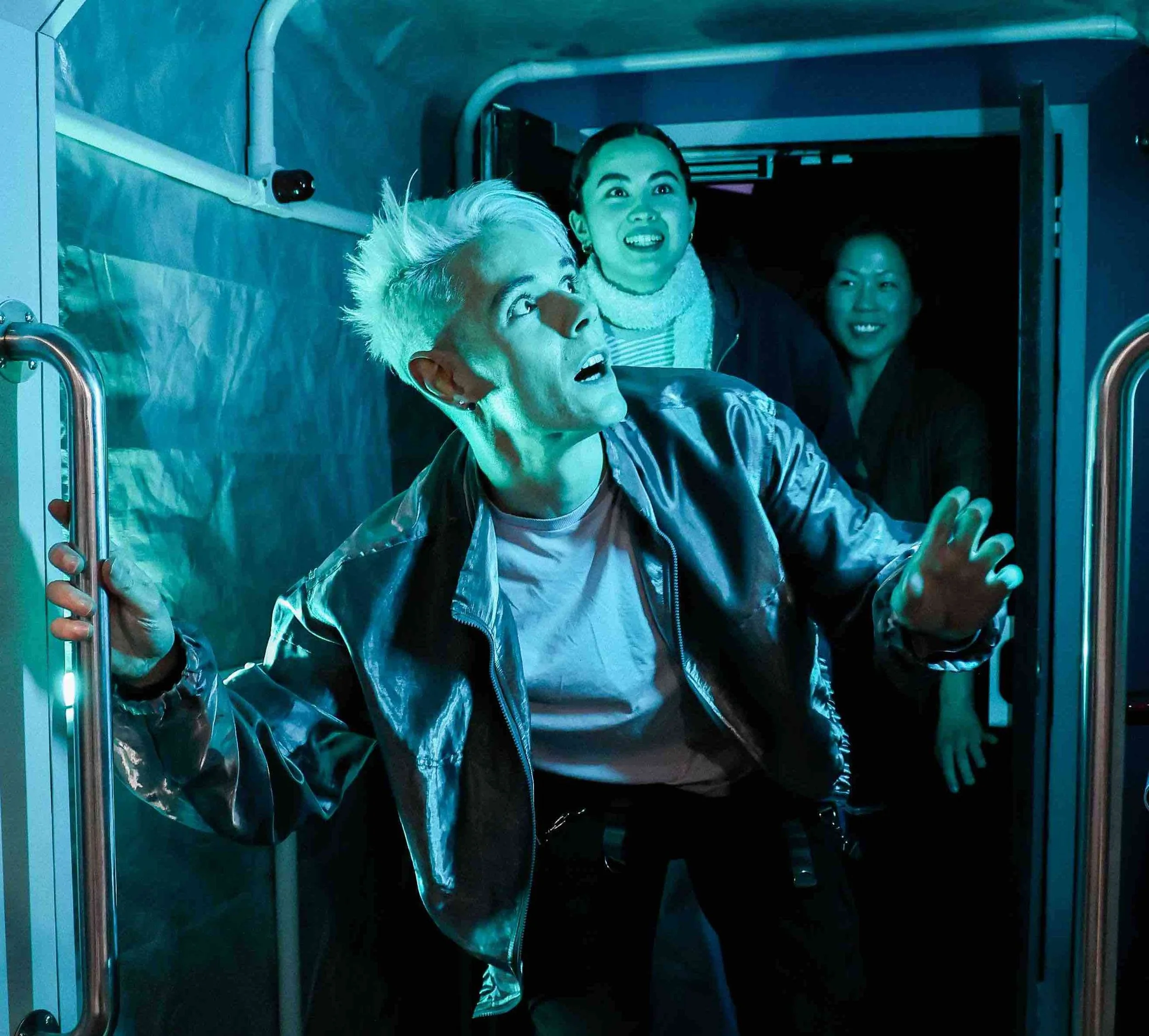 People in an elevator, looking upward with expressions of surprise or curiosity, illuminated by colorful lighting.