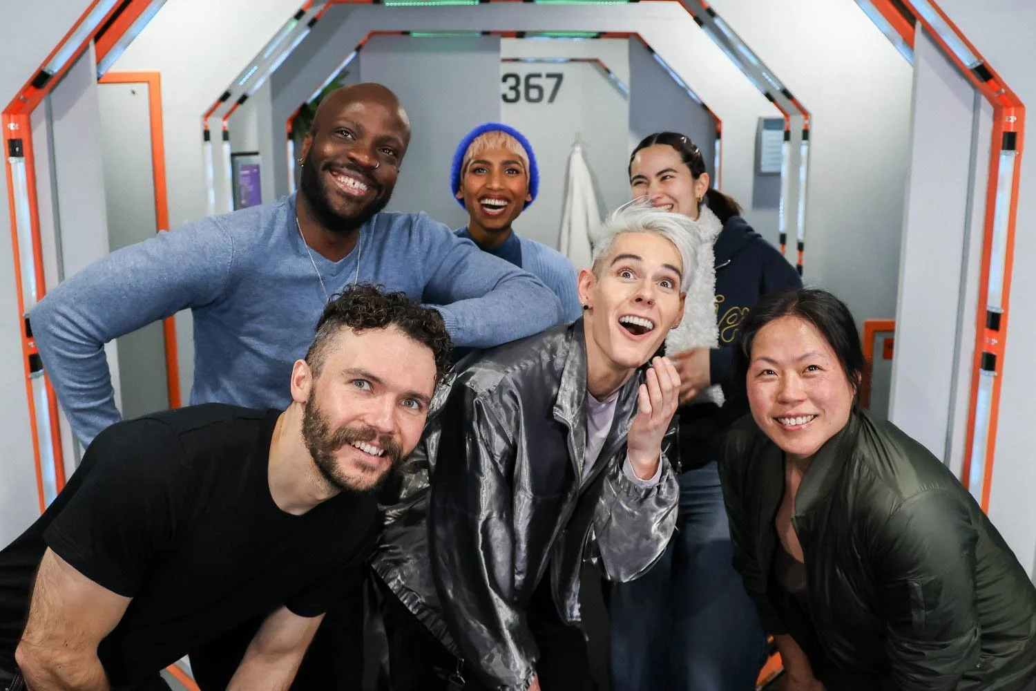 A diverse group of seven smiling friends in a futuristic hallway with orange and gray accents.