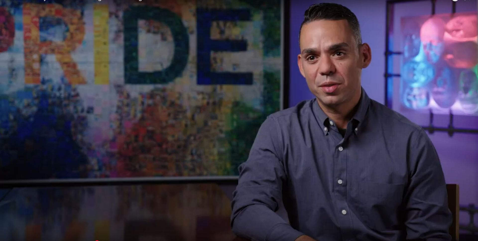 A man sitting against a blue and purple background giving an interview