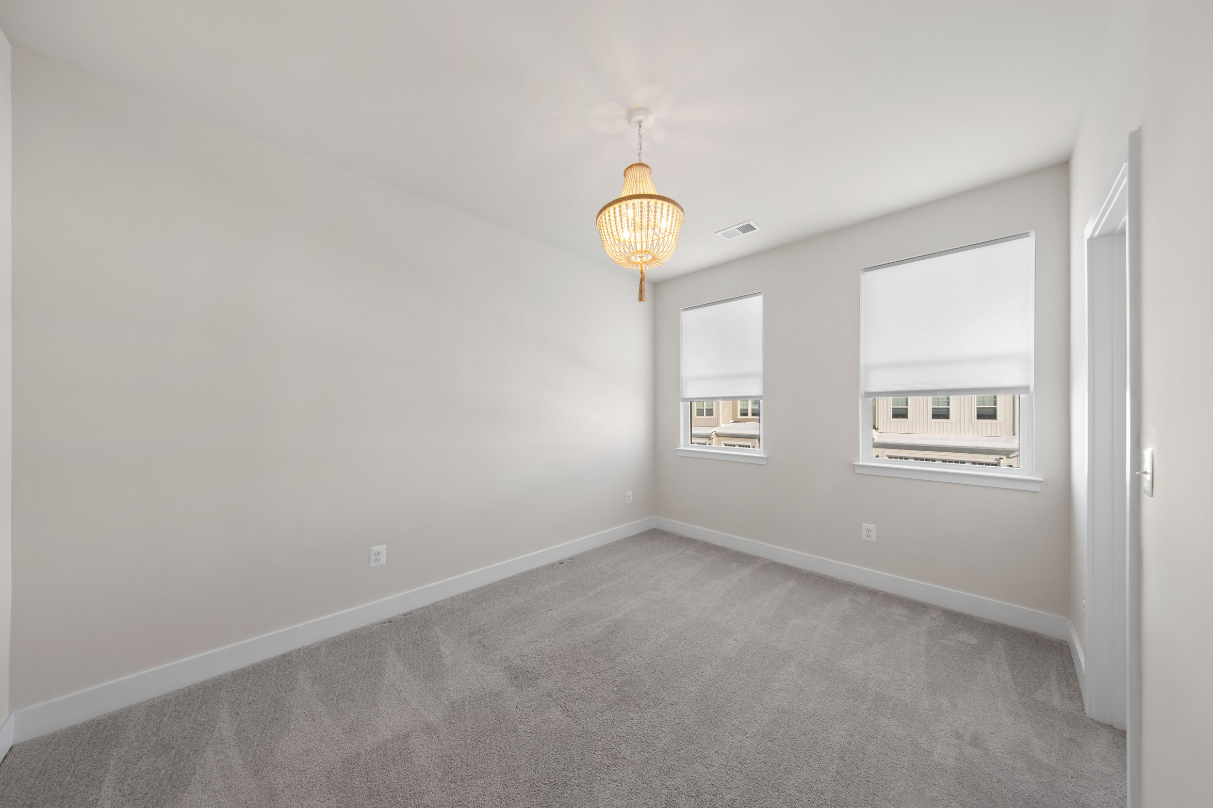 Empty room with beige carpet, white walls, two windows with rolled shades, a hanging light fixture, and a partially open door.