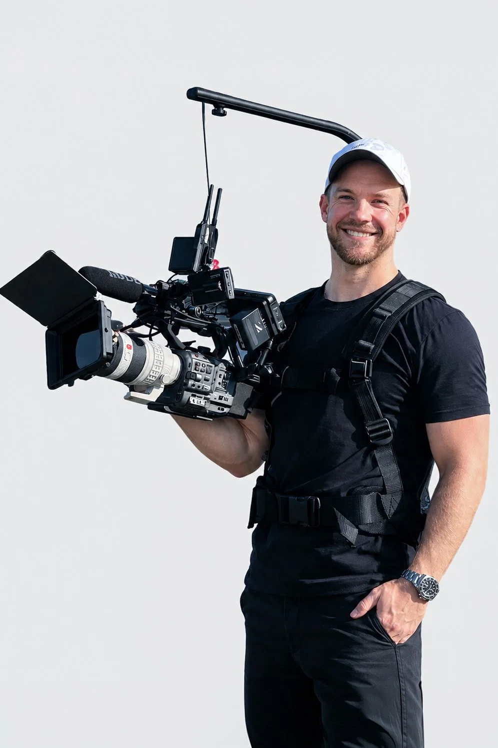 A man with a beard and short hair standing in a living room, holding a camera on a tripod, smiling, with a sofa, paintings of seashells, lamps, and pillows in the background.