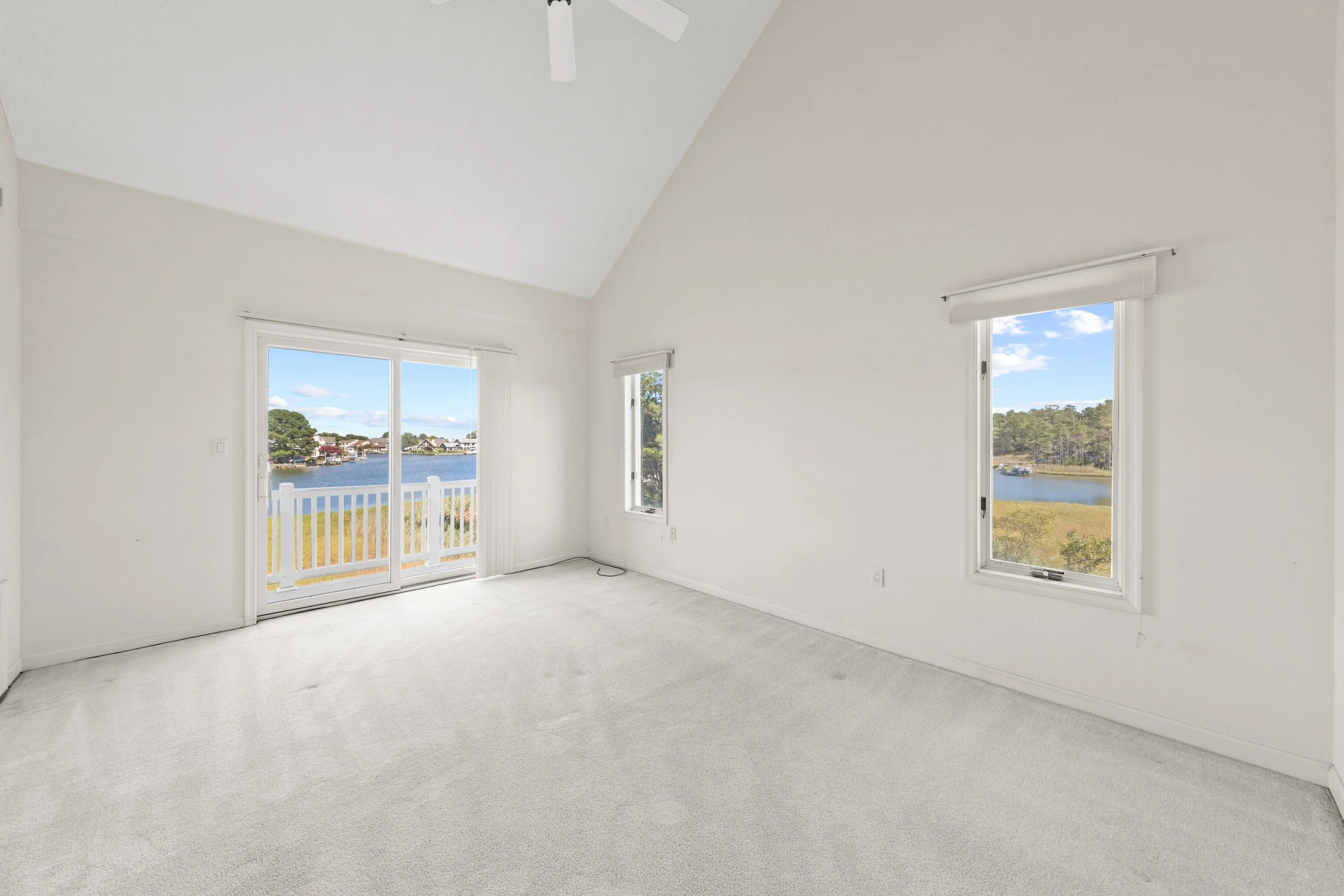 Empty room with white walls, beige carpet, two windows, and a sliding glass door leading to a balcony with a view of a river and greenery.