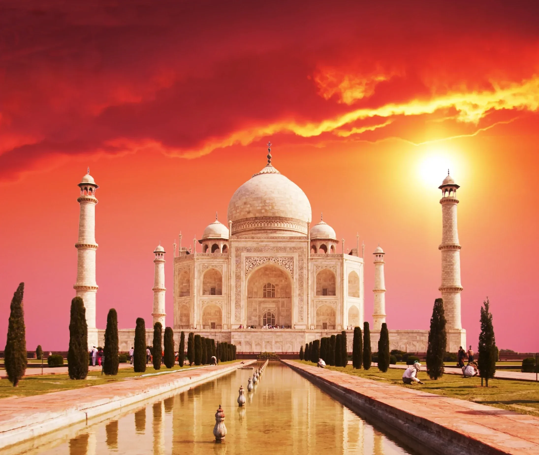 The Taj Mahal at sunset with a vibrant orange sky, reflecting in the long pool in front of the monument, surrounded by visitors and trees.