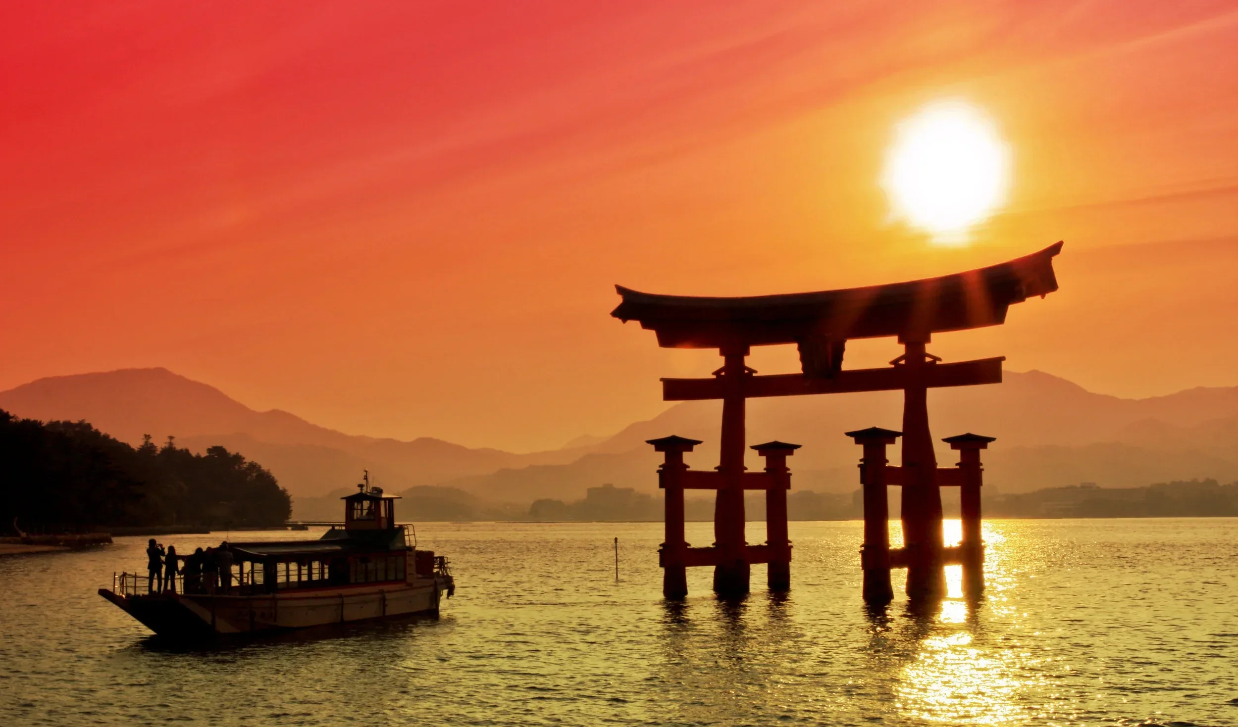 A traditional Japanese torii gate standing in the water at sunset, with a boat and people nearby, mountains in the background, and the sky glowing in shades of orange and pink.