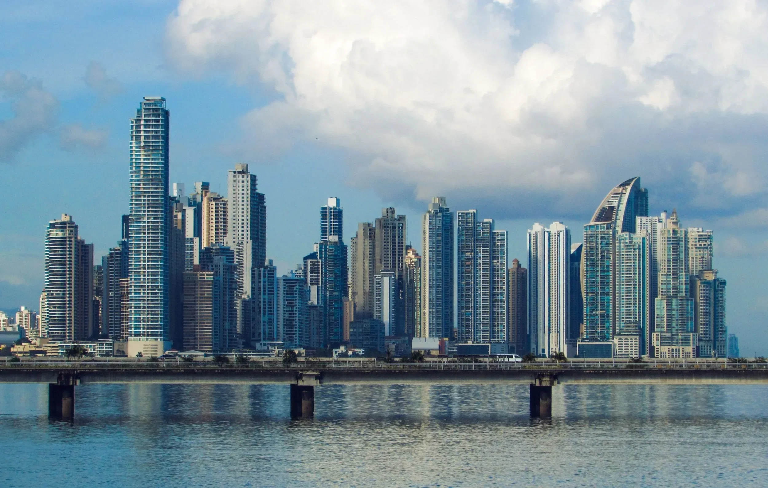 City skyline with numerous modern high-rise buildings and a distinctive curved skyscraper, viewed across a river with a bridge in the foreground and a partly cloudy sky overhead.