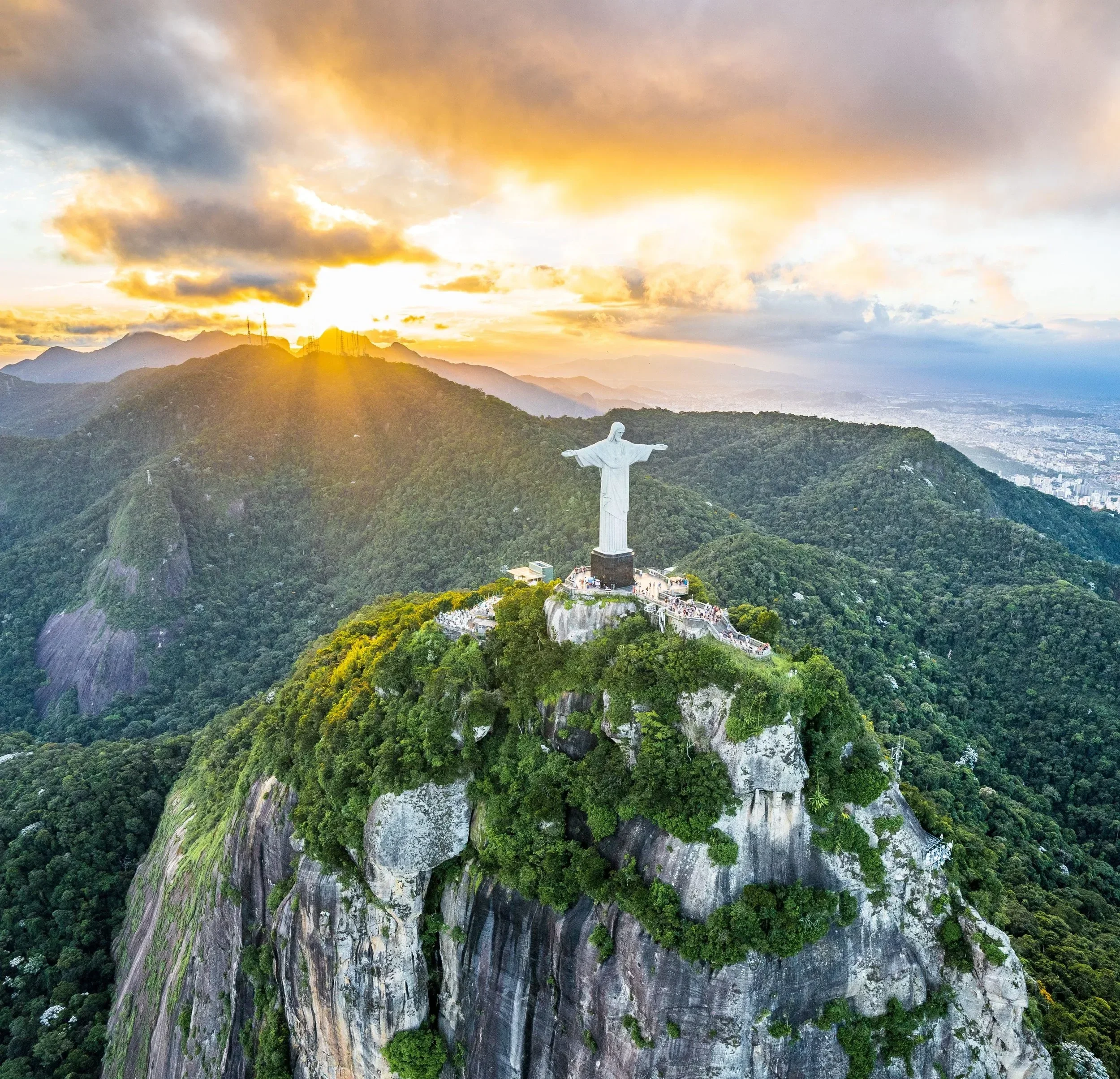 Christ the Redeemer statue overlooking Rio de Janeiro at sunset with a mountain range, cloudy sky, and cityscape in the background.