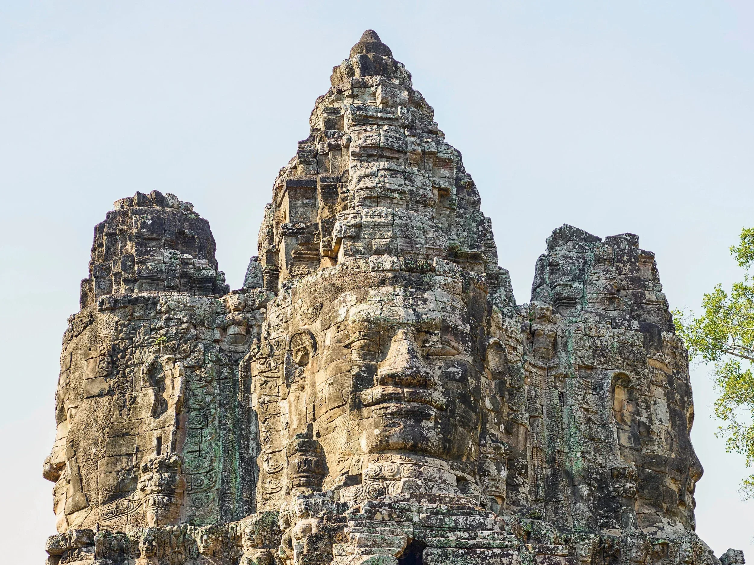 Ancient stone temple tower with detailed carvings and weathered surfaces, set against a pale sky with some green foliage visible at the bottom right.