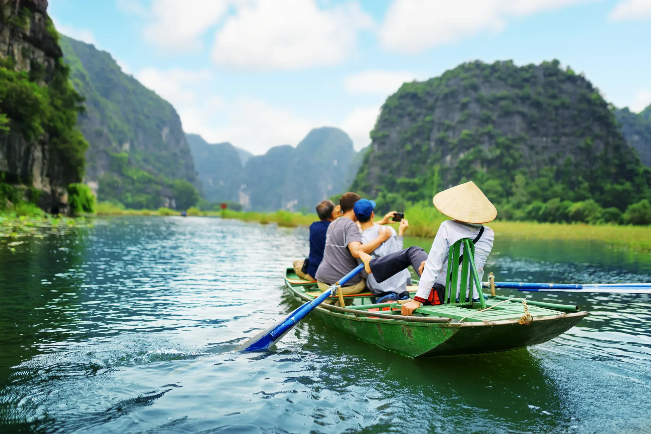 A group of people on a bamboo raft, with one person wearing a traditional conical hat, rowing on a river surrounded by lush green mountains under a cloudy sky.