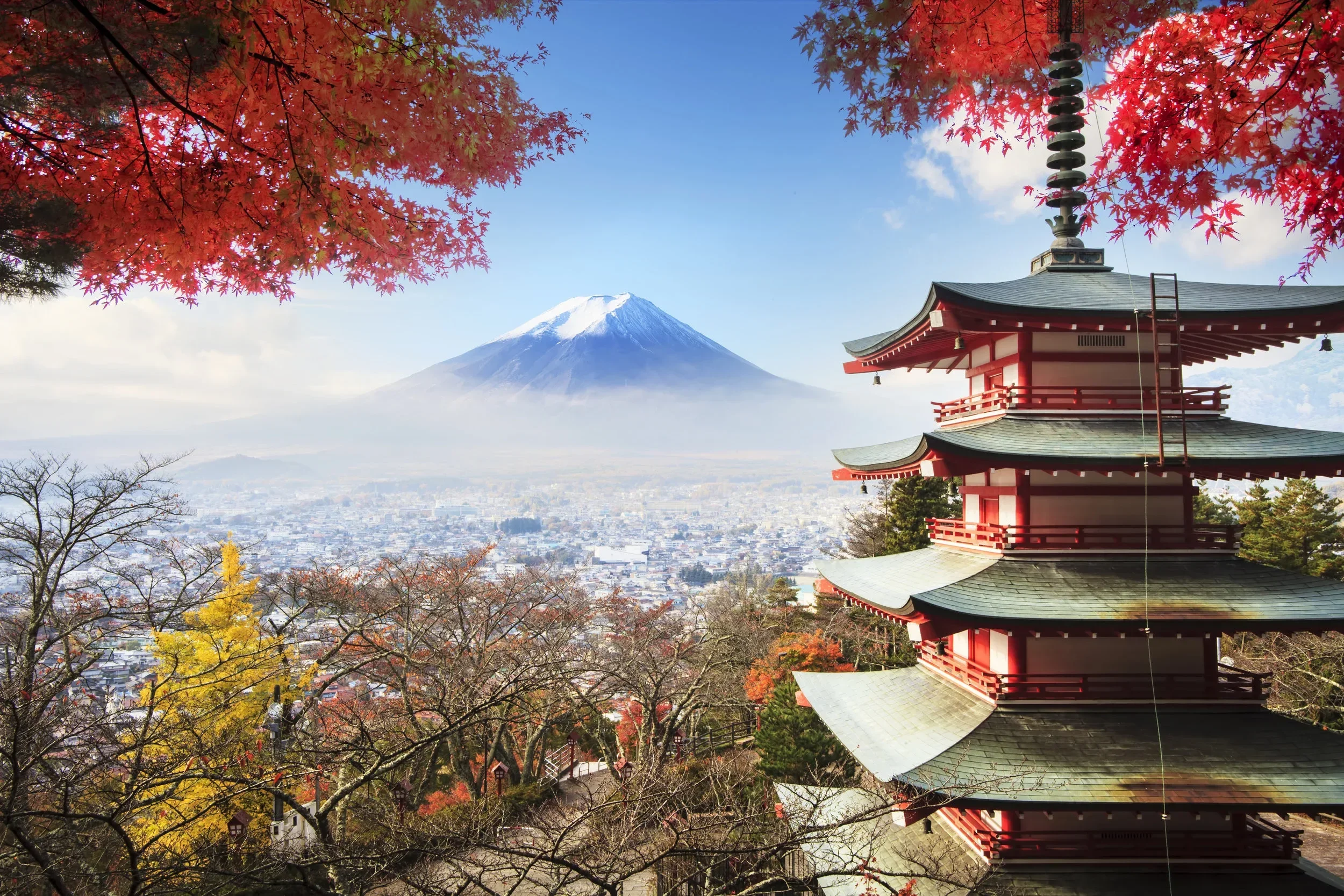 A traditional red pagoda style building with curved roofs under autumn trees with red, yellow, and orange leaves, Mount Fuji with snow on its peak in the background, and a blue sky with some clouds.