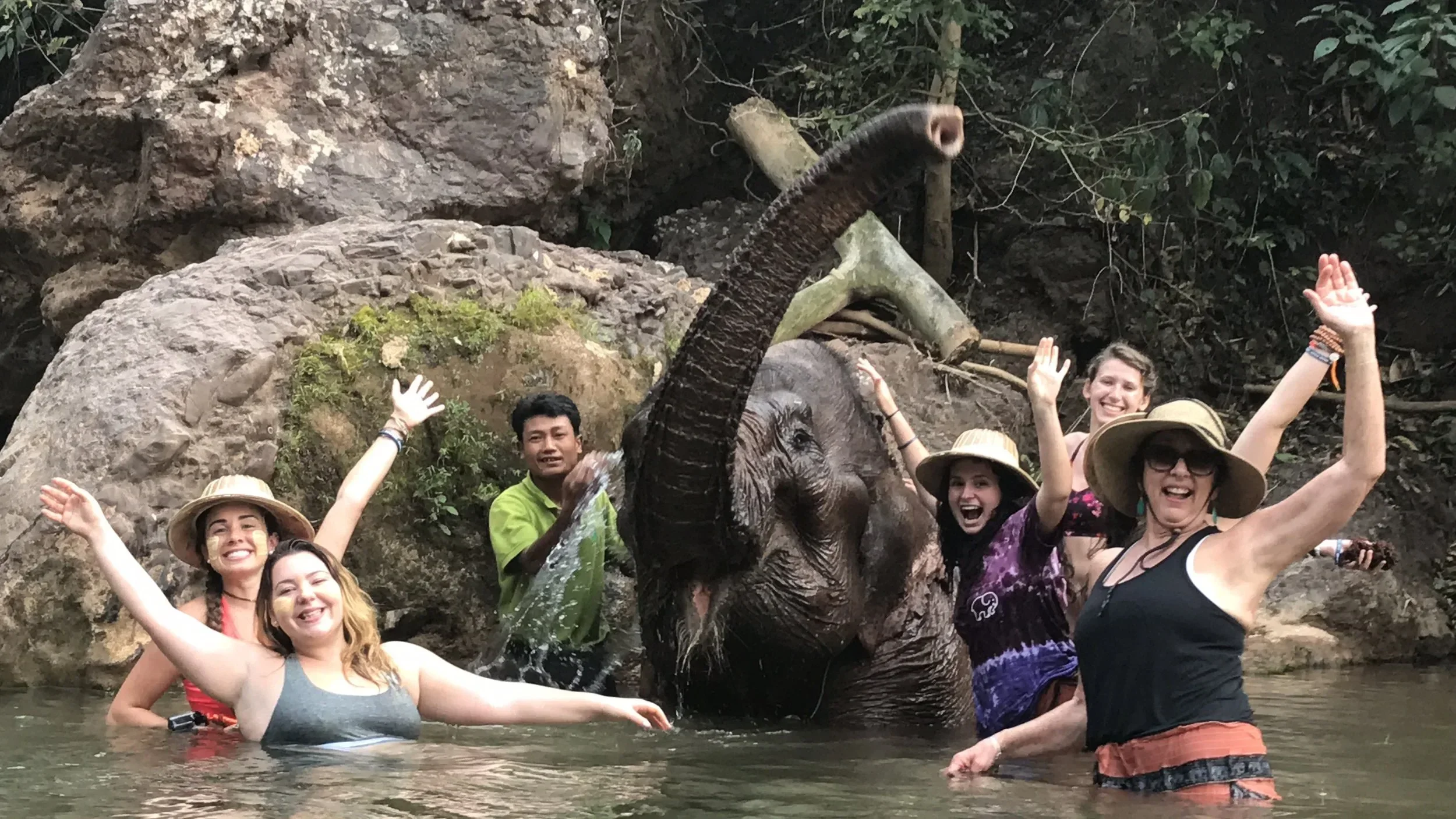 Group of smiling people in water with a small elephant statue or sculpture and large rocks in the background.