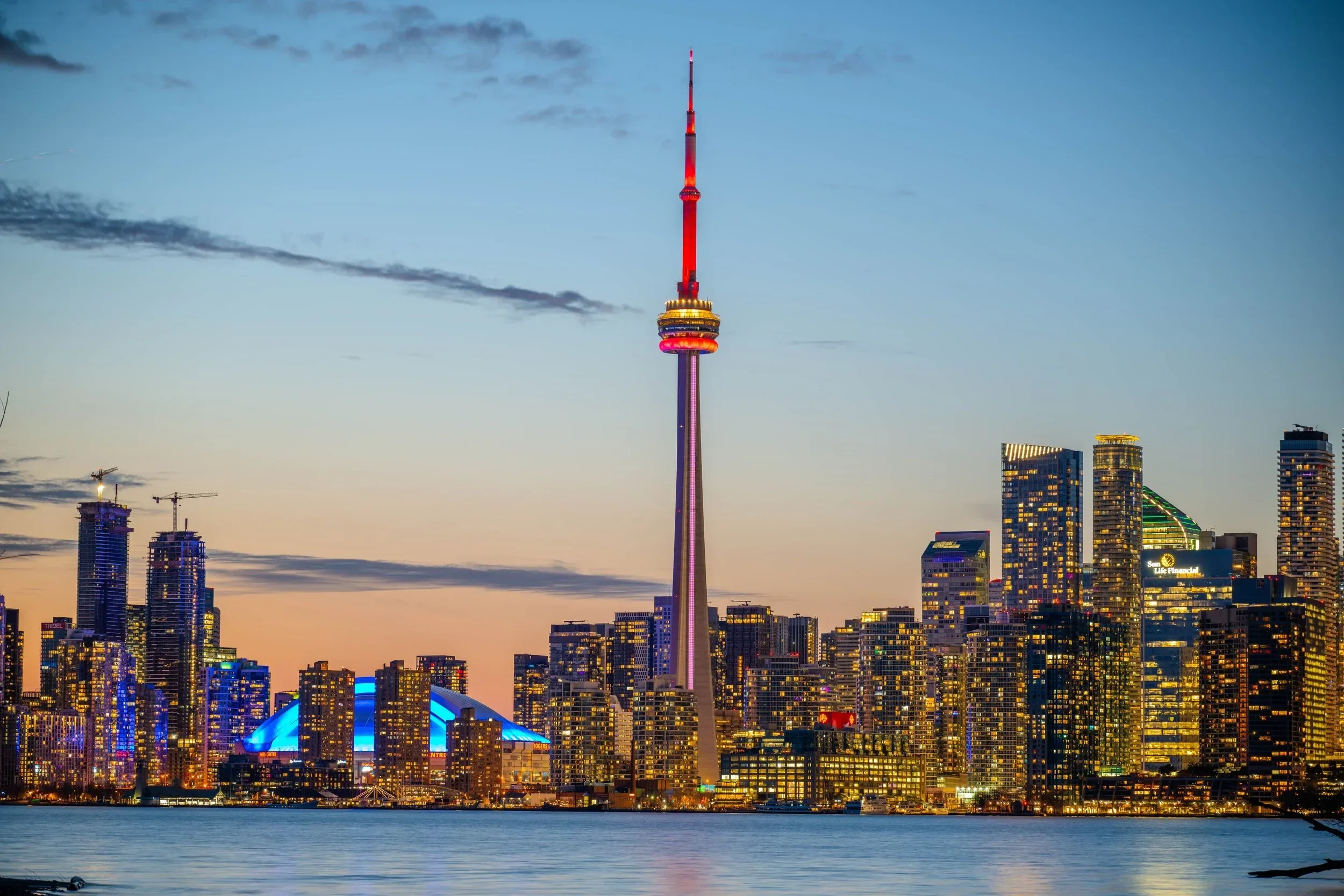 Nighttime view of Toronto skyline with the CN Tower illuminated in red, yellow, and purple lights, waterfront in the foreground, and skyscrapers with colorful lights.