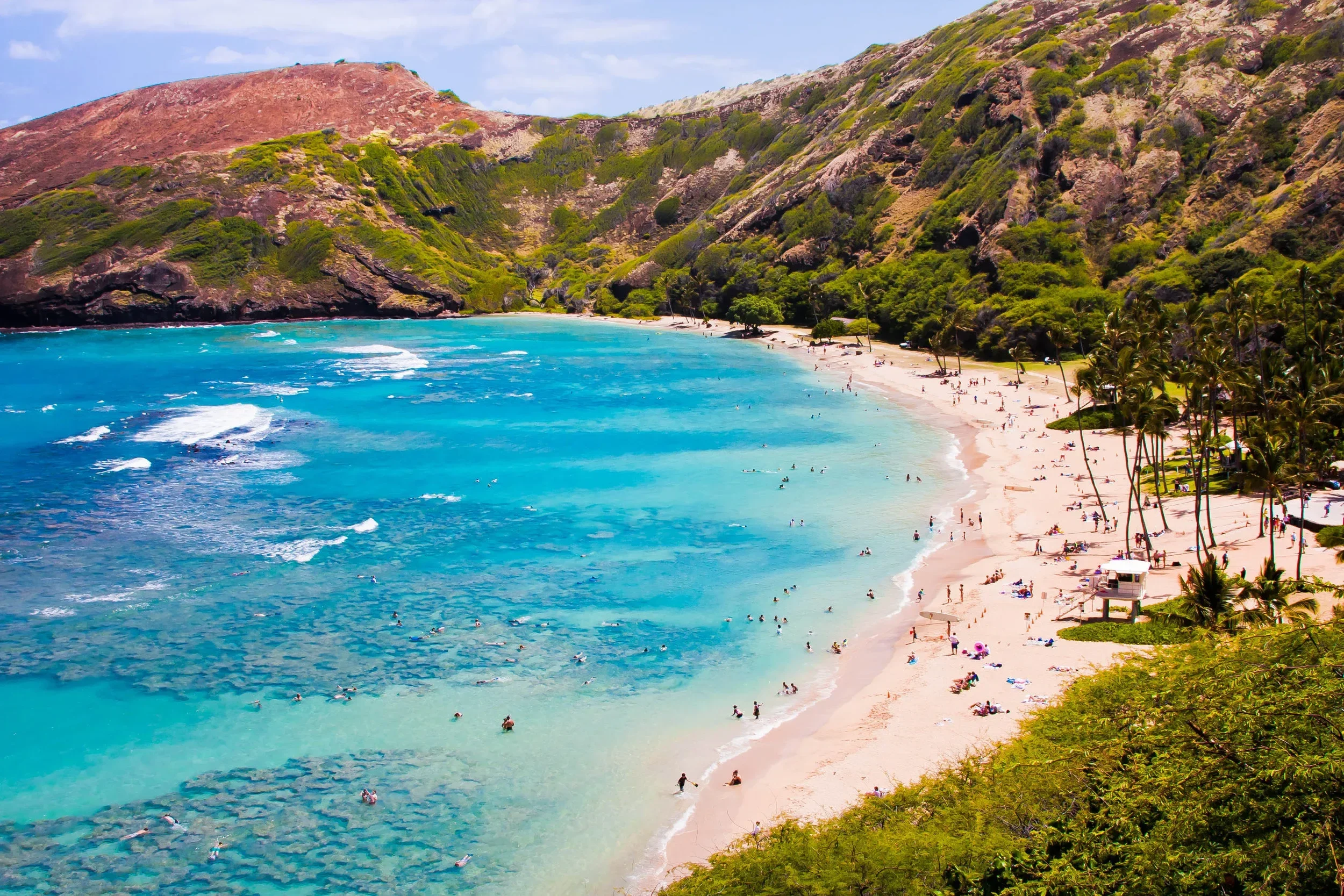 A tropical beach with turquoise water, white sand, lush green trees, and steep mountains in the background. People are swimming, relaxing, and enjoying the scenery.
