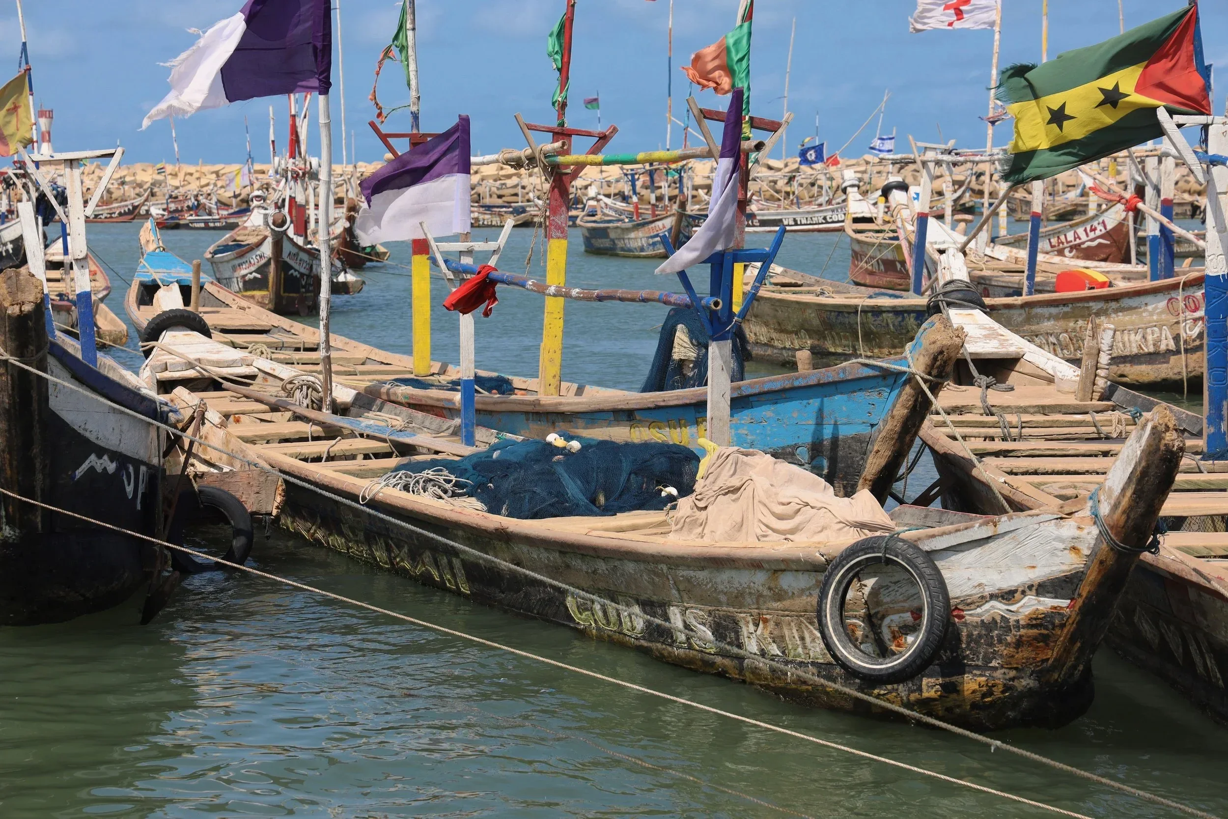 A harbor filled with colorful small fishing boats with flags, tied together and floating on water, with a rocky breakwater in the background and a clear blue sky overhead.