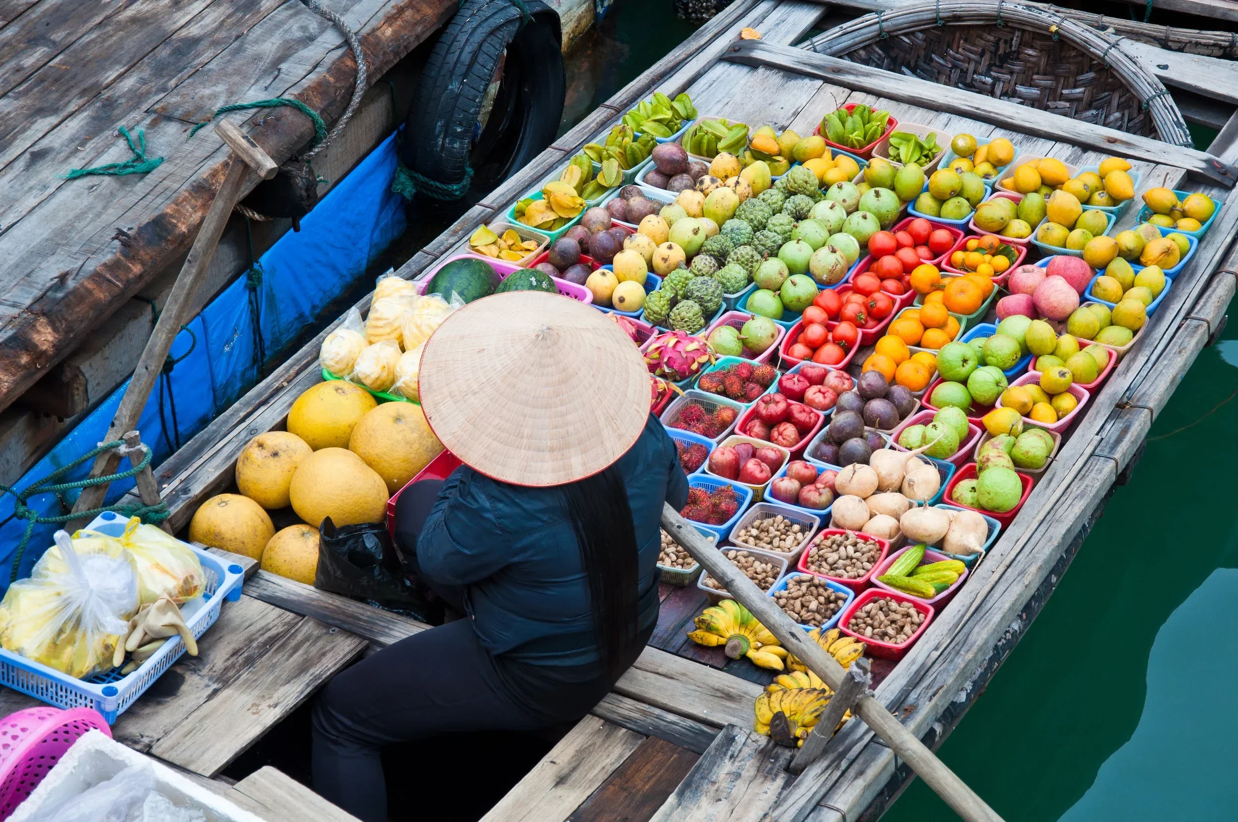 A wooden boat floating on water filled with various fruits and vegetables in colorful containers, with a person wearing a traditional conical hat and dark clothing sitting inside.