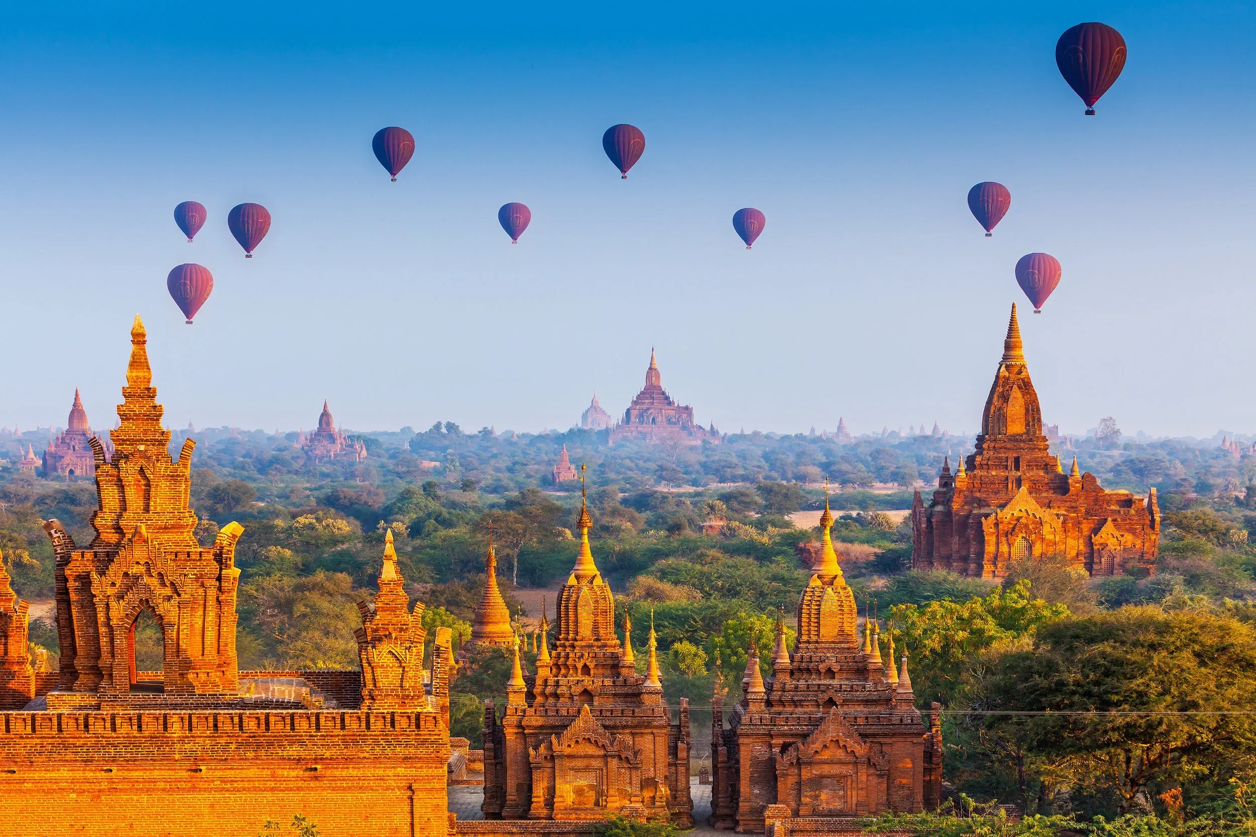 Multiple hot air balloons floating over ancient temples and pagodas in a landscape with greenery and distant ruins under a clear blue sky.