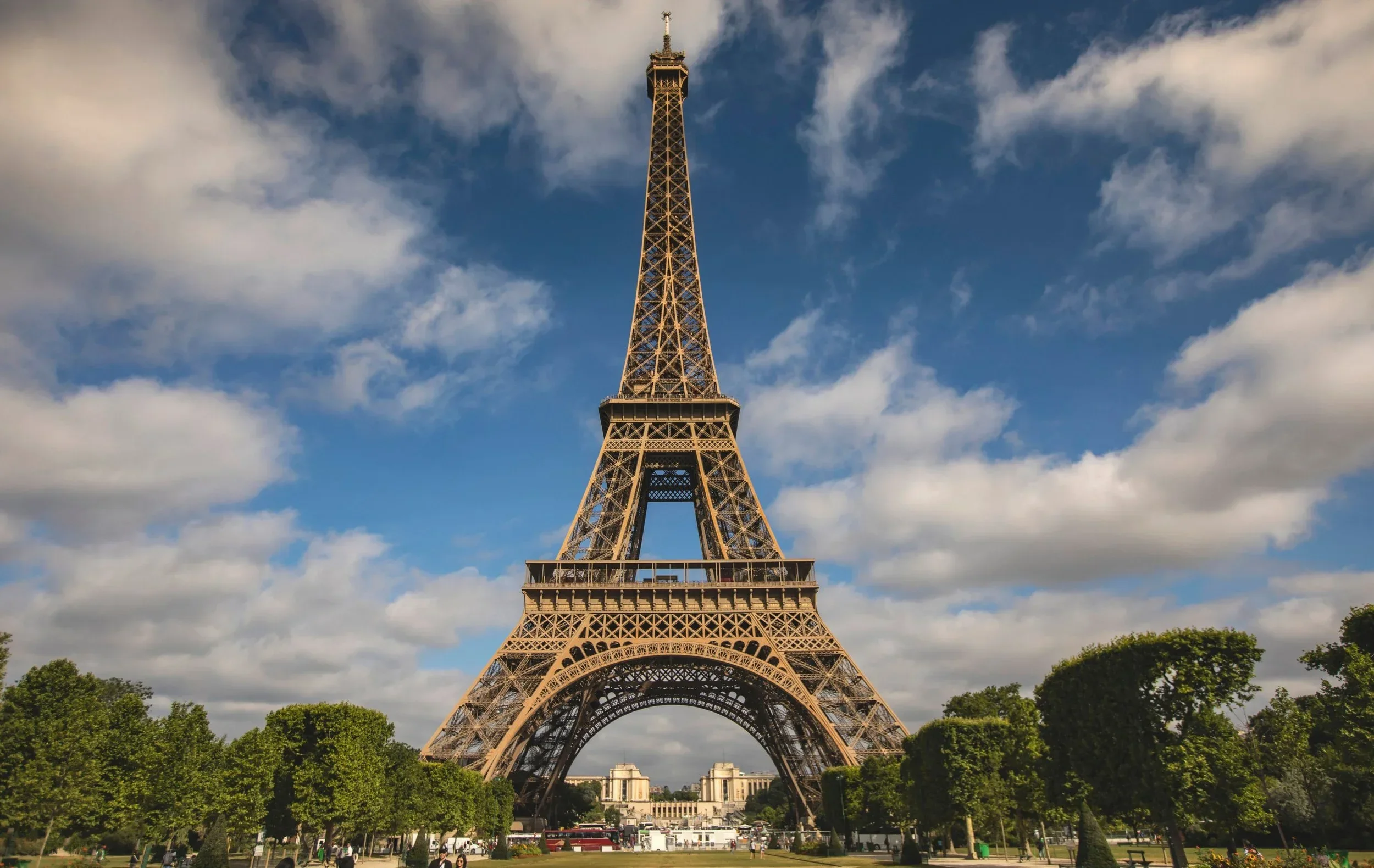 The Eiffel Tower in Paris, France, seen from the ground with a partly cloudy sky in the background and green trees at the base.