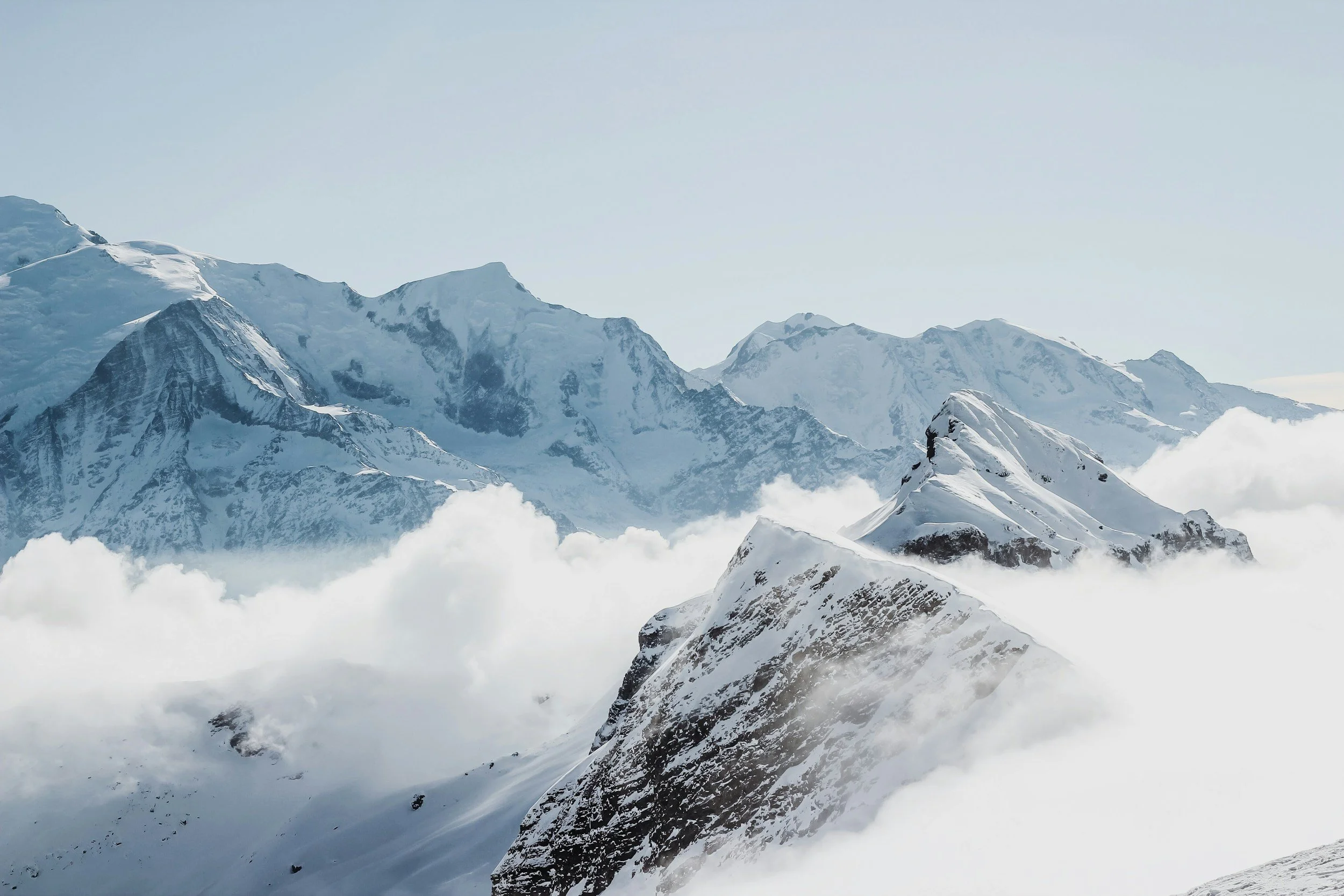 Snow-covered mountain peaks with clouds surrounding them against a clear sky.