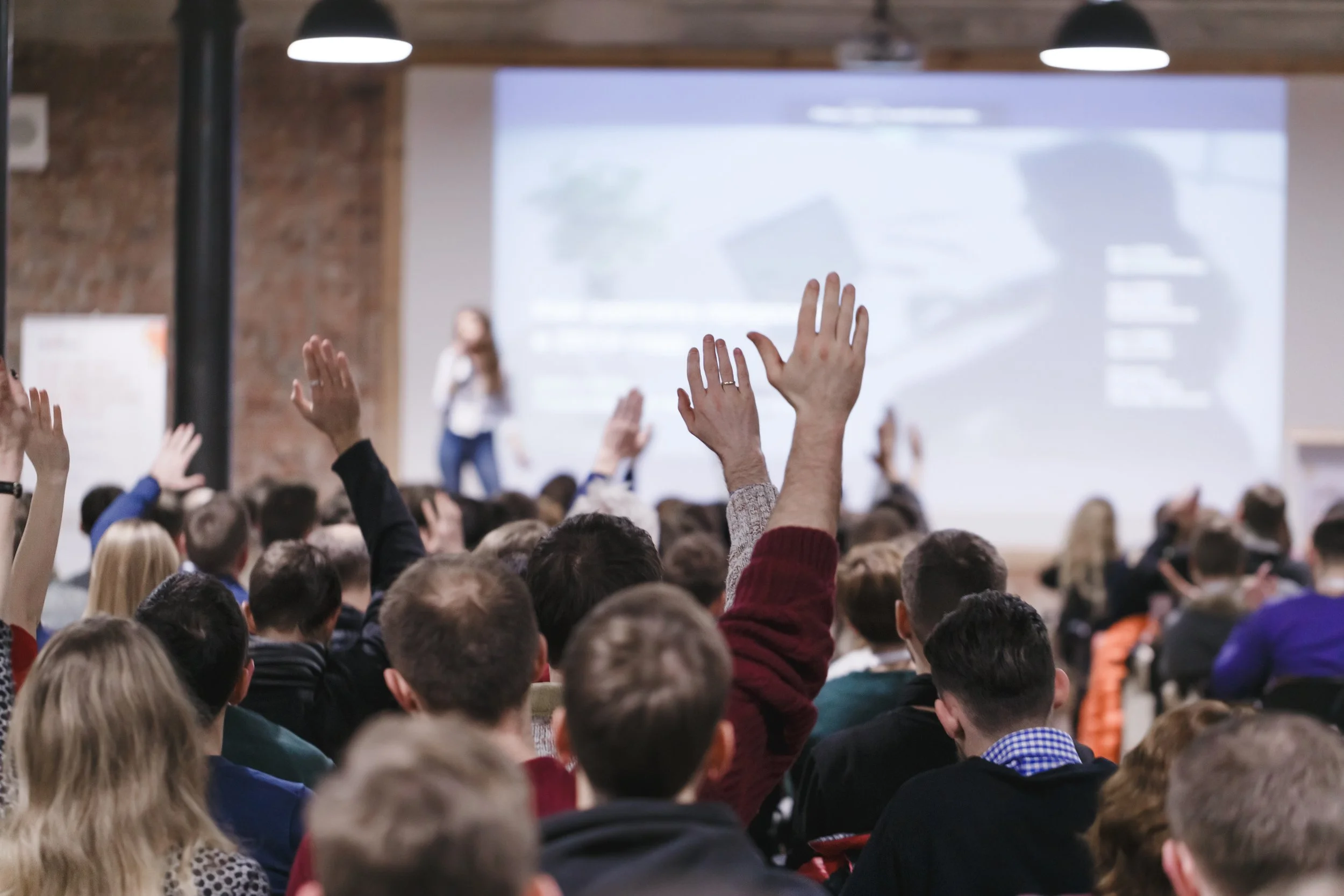 Audience at a conference or seminar with many people raising their hands, facing a stage with a presenter and a large screen.