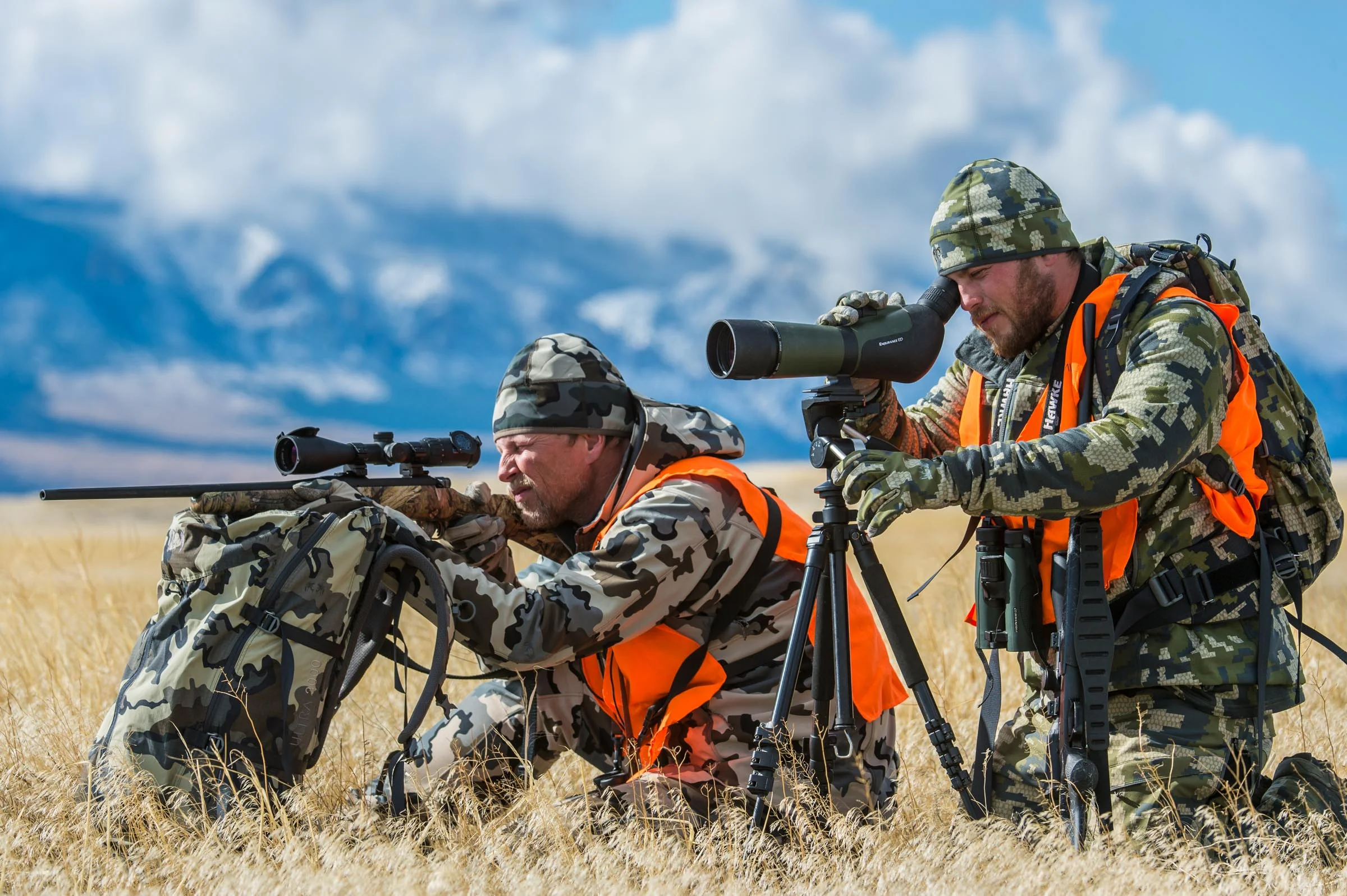 Two hunters one looking through a spotting scope one through a rifle aim to hunt deer on the Montana prairie in the shadow of the Rocky Mountains. © Tony Bynum