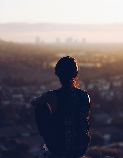 The silhouette of a young woman sitting on a rock and looking out over a rocky landscape and a distant city. See how far you can stretch all of your senses in every direction. What can you hear, see, smell, taste, feel?