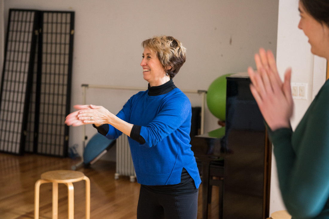 Belinda Mello teaches the Alexander Technique to a group of actors. She rubs her hands together and smiles teaching her students about the use of energy. A student smiles in the right side of the picture.