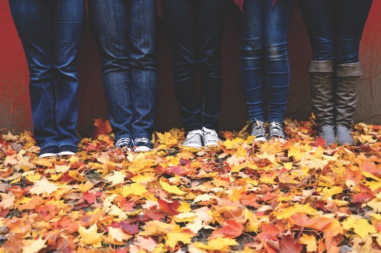4 young people stand against a wall in jeans and sneakers. It’s fall and there’s leaves on the ground. They’re doing an Alexander Technique etude where they see if they can do less while sliding down the wall.