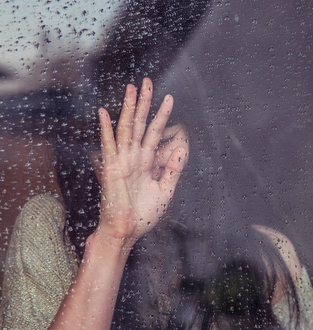 woman with long hair rests her hand on the glass of a window, her eyes are shut. There are drops of rain on the outside of the window.