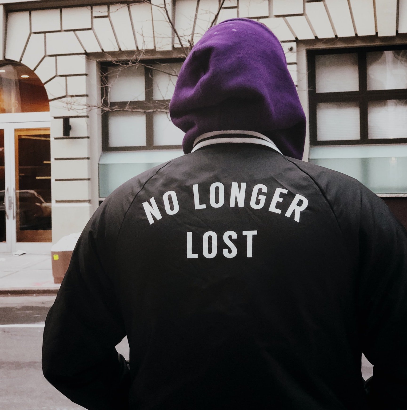 The back of a person on a city street. Letters on the back of their black jacket read, No Longer Lost