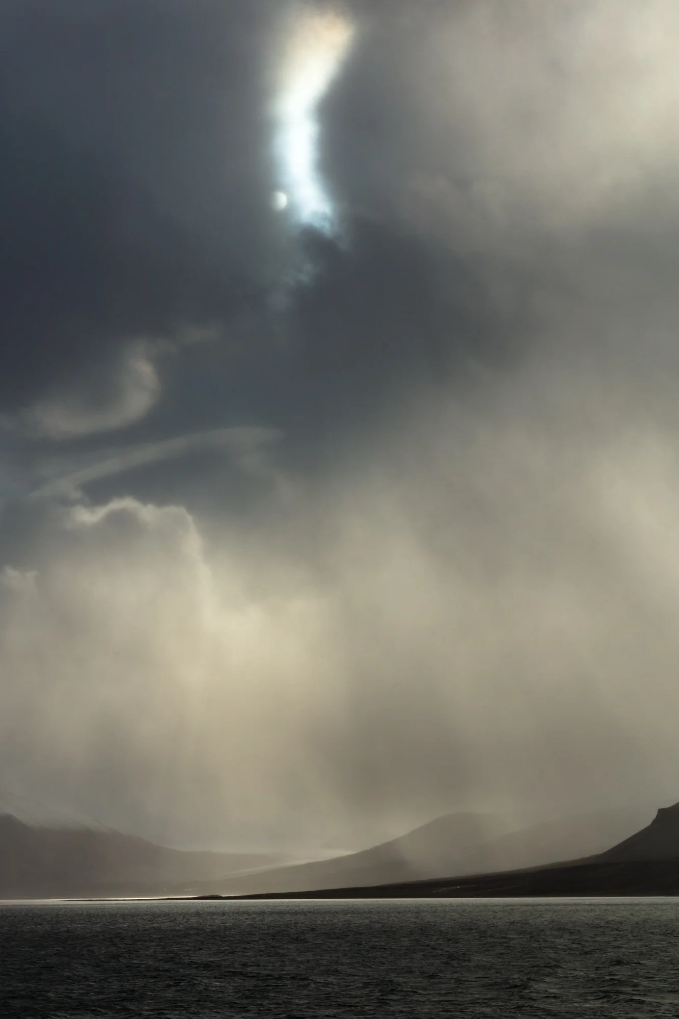 The soft arctic sun peers along the edge of a storm-obscured lenticular cloud formation, illuminating it with a silver bolt of light above the shadowy tundra below. The morning began dark and gloomy with heavy winds, fog, and rain that hid the fjords behind heavy weather. As the storm cleared for a few brief moments, the clouds, landscape, and sea were bathed in sepia-gold rays of light.
