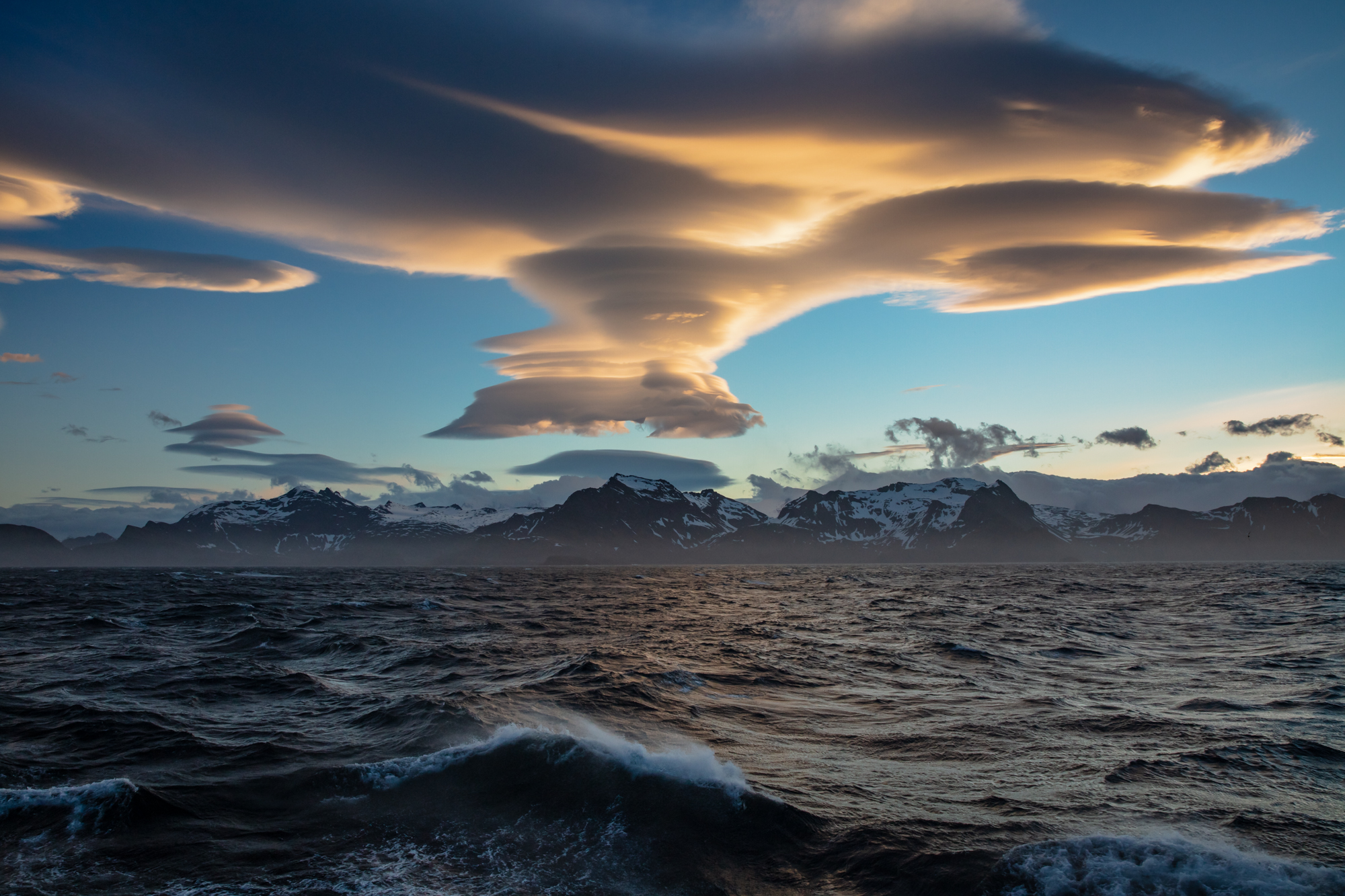 A lenticular cloud formation sits like a series of flying saucers above the highest peaks of South Georgia that rise from the wind-whisked seas below. As the ship-clock neared 11PM , one of the expedition crew casually called out on the intercom that there were lenticular clouds on the horizon if anyone was still awake. After a long day of image making onshore while fighting a wicked wind, I had to muster all of my strength to bundle up and get out on deck. Though the promise of lenticular clouds is enough to get any photographer out of bed. Though I have seen lenticular clouds before, I have only seen singular versions of the strange saucer shaped clouds. When I stepped outside to see a rare sky full of lenticular clouds clustered atop one another like a stack of golden pancakes, I froze in disbelief of the beauty. As a wall of mountains in the middle of one of the windiest places on earth, South Georgia the perfect storm for lenticulars. Clouds like these only form when intense winds - which neared hurricane speeds that day - are forced into a standing wave by mountains, leaving otherworldly disks above the highest summits.