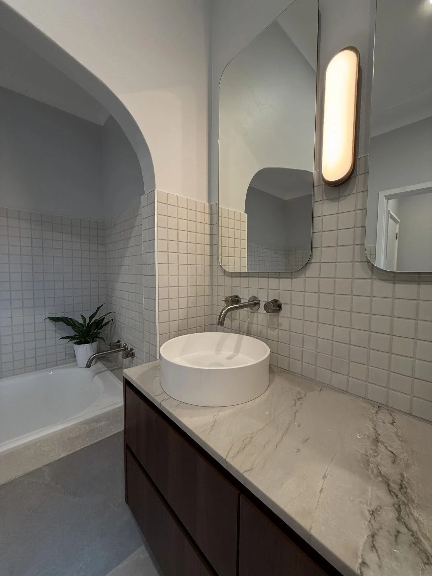Modern bathroom with a marble countertop, round white vessel sink, wall-mounted faucet, rectangular mirror, and a tiled wall with a potted plant near the bathtub.