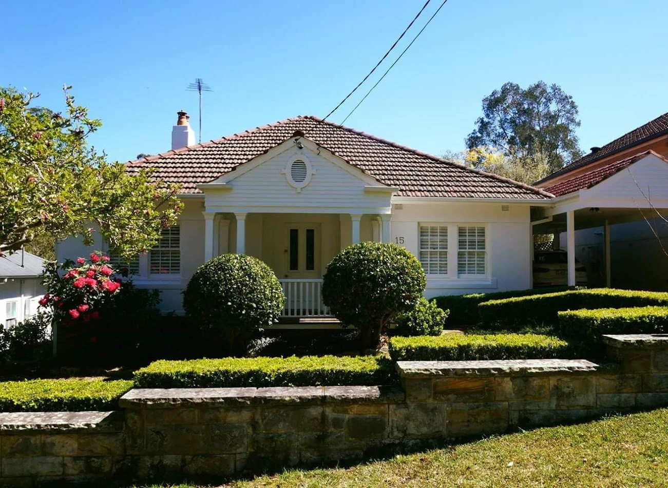 A white single-story house with a red-tiled roof, front porch, and well-maintained garden with trimmed bushes and a stone retaining wall, under a clear blue sky.