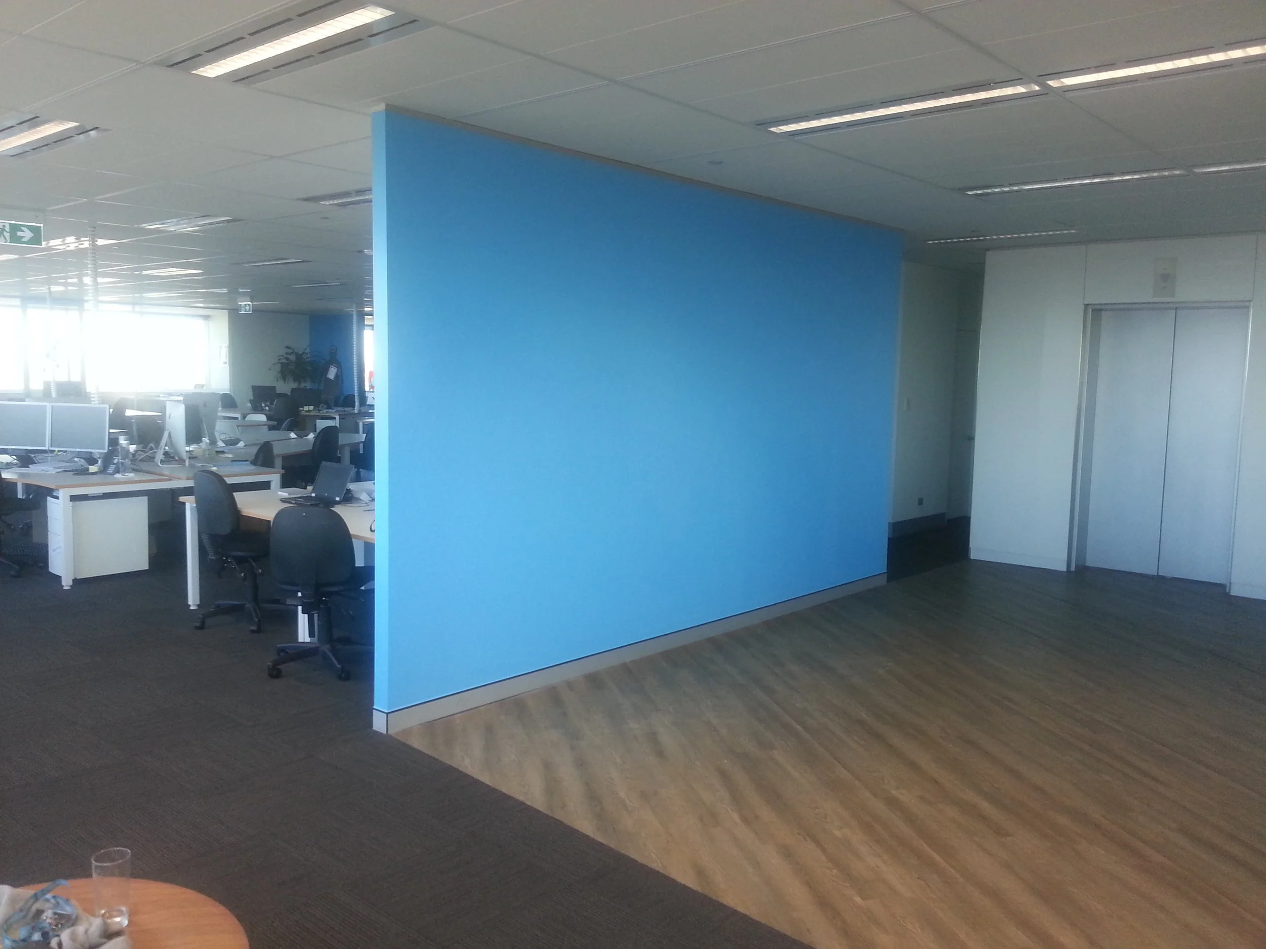 Office space with multiple desks, chairs, and computer monitors, divided into two sections by a blue partition wall. The area has large windows allowing natural light.
