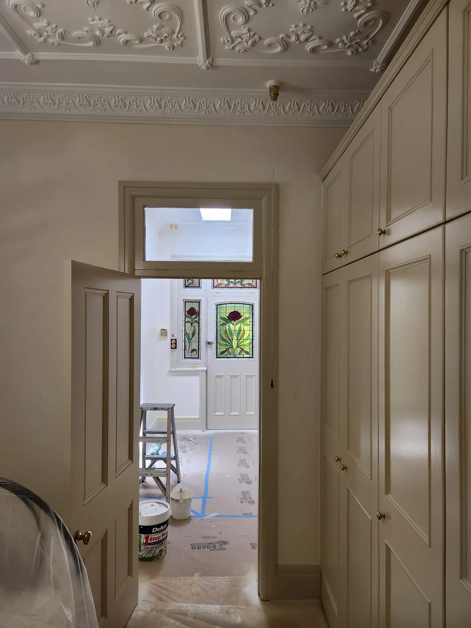 View through a doorway into a hallway under renovation, with decorative stained glass windows featuring floral designs, construction tools, and materials on the floor.