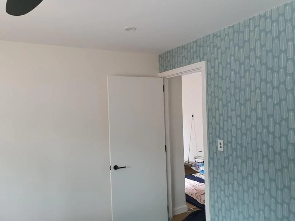 A room with white walls and a patterned blue accent wall, partially open door leading to another room, and a small portion of a bed visible in the adjacent room.