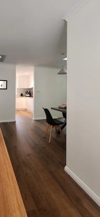 Interior view of a modern home with white walls, wooden flooring, a black dining chair, and a dining table with some items on it, leading to a kitchen area.
