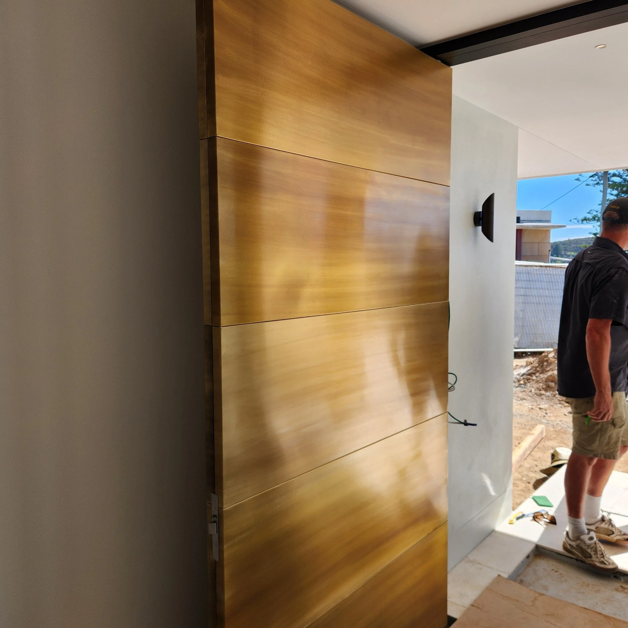 Interior view of a house under construction, showing a wooden panel wall, a partially visible man wearing shorts and a black shirt, construction tools on the floor, and an outdoor view through the opening.