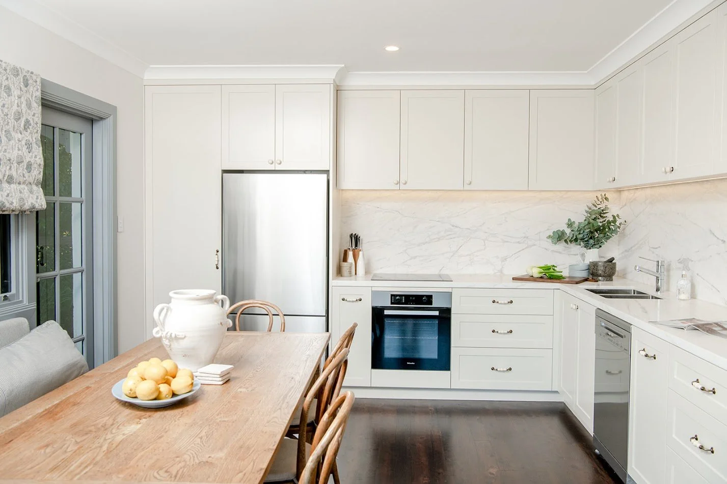 Bright kitchen with white cabinets, marble backsplash, stainless steel appliances, wooden dining table, and natural light.