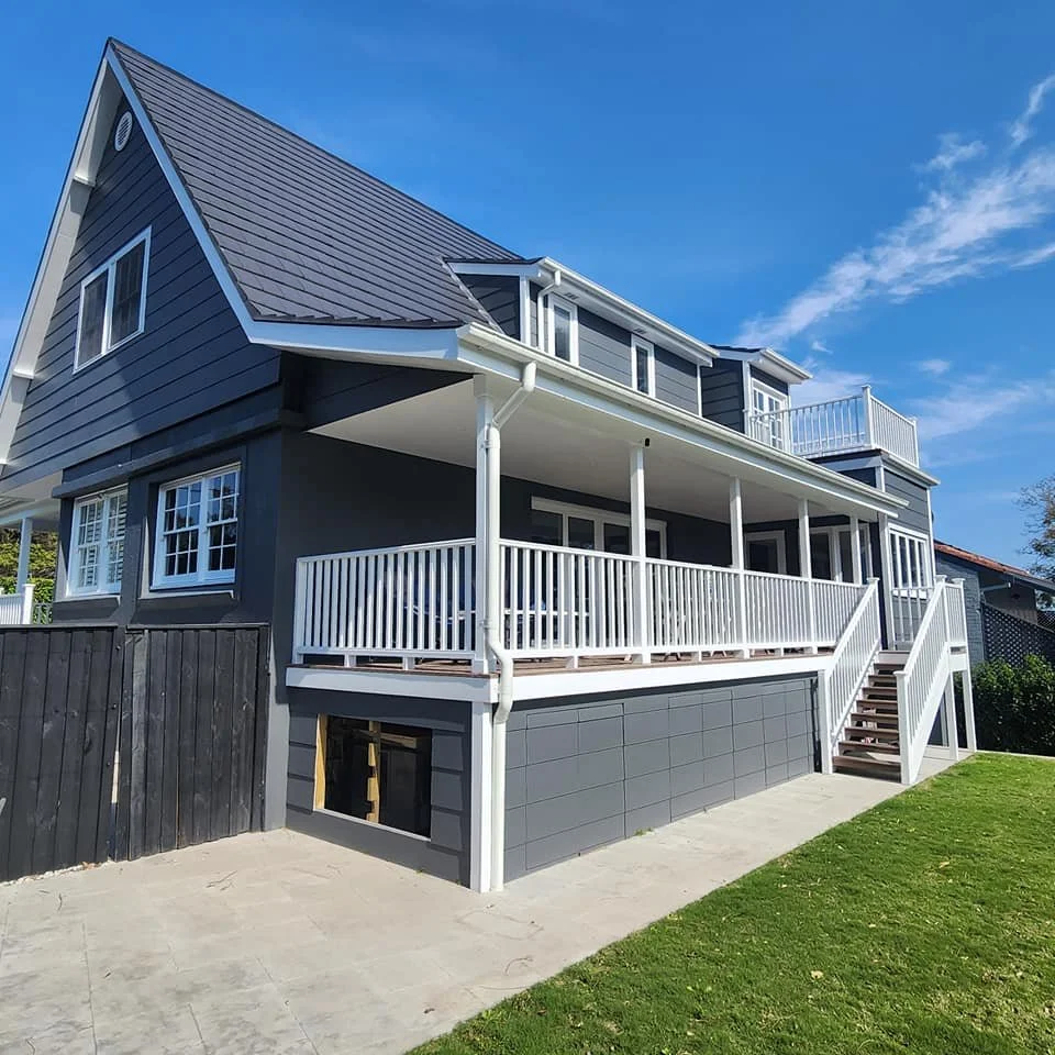 A modern two-story house with a dark blue exterior and white trim, featuring a large front porch with white railings, stairs leading up, and multiple windows, under a clear blue sky.